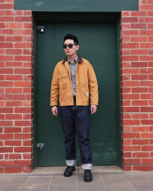 A man wearing a Maden Canvas golden-tan Chore coat, blue denim jeans, a gray flannel shirt, and black sunglasses stands in front of a dark green door framed by a red brick wall.