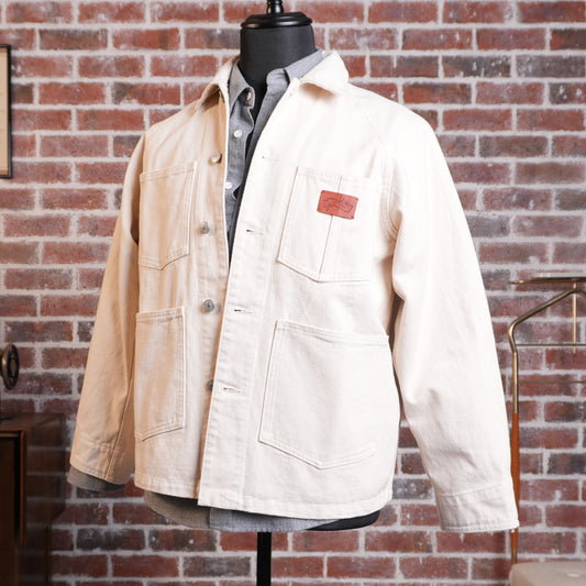 A natural ecru cotton canvas utility jacket with a brown leather patch on the chest pocket, displayed on a black mannequin torso against a red brick wall background.