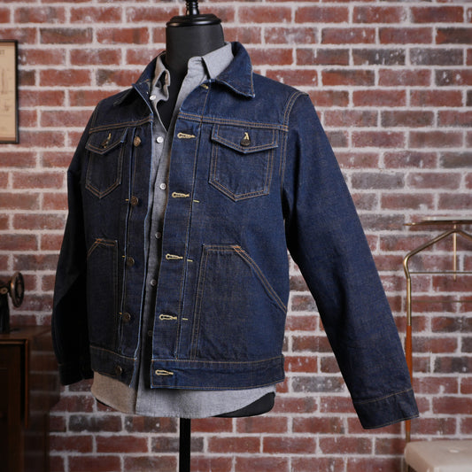 A dark blue denim jacket displayed on a black mannequin torso, layered over a light gray button-up shirt. The jacket features classic styling with front flap pockets, button closures, and contrast stitching, set against a rustic red brick wall background.