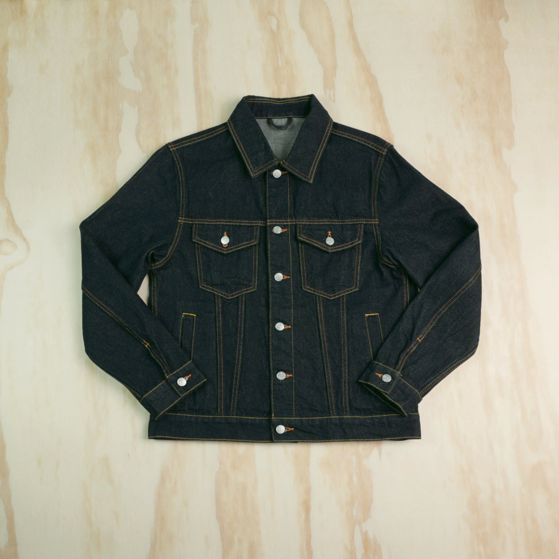 Dark indigo Type III selvedge denim trucker jacket with orange stitching laid flat on a wooden surface.