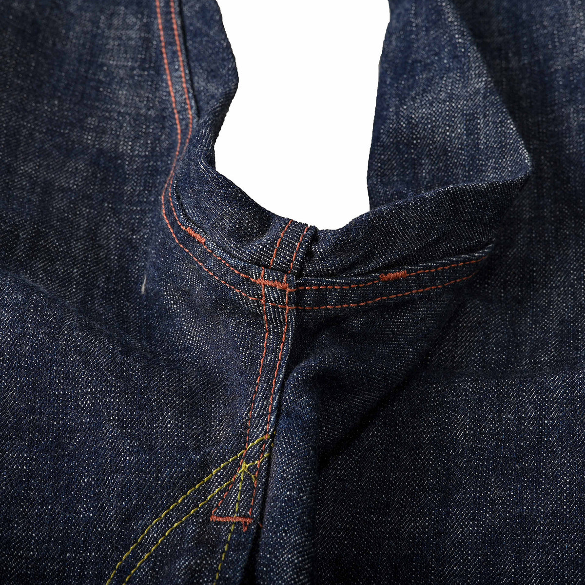 Close-up of dark indigo denim fabric showing the waistband and center seam with contrasting orange and yellow stitching. Shows bartack reinforcement stitching at 3 points on the crotch seam area.