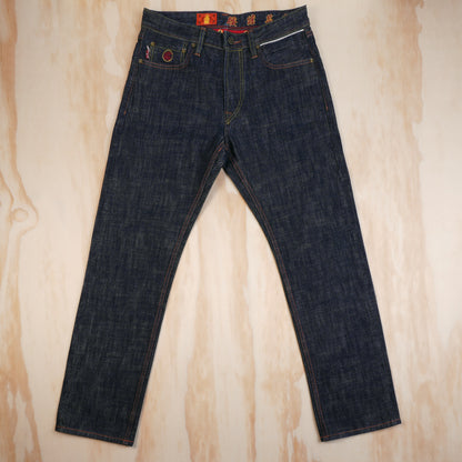 Dark indigo blue selvedge denim jeans laid flat on a light wood grain background, featuring a red and yellow waistband label and a red circular patch on the front pocket.
