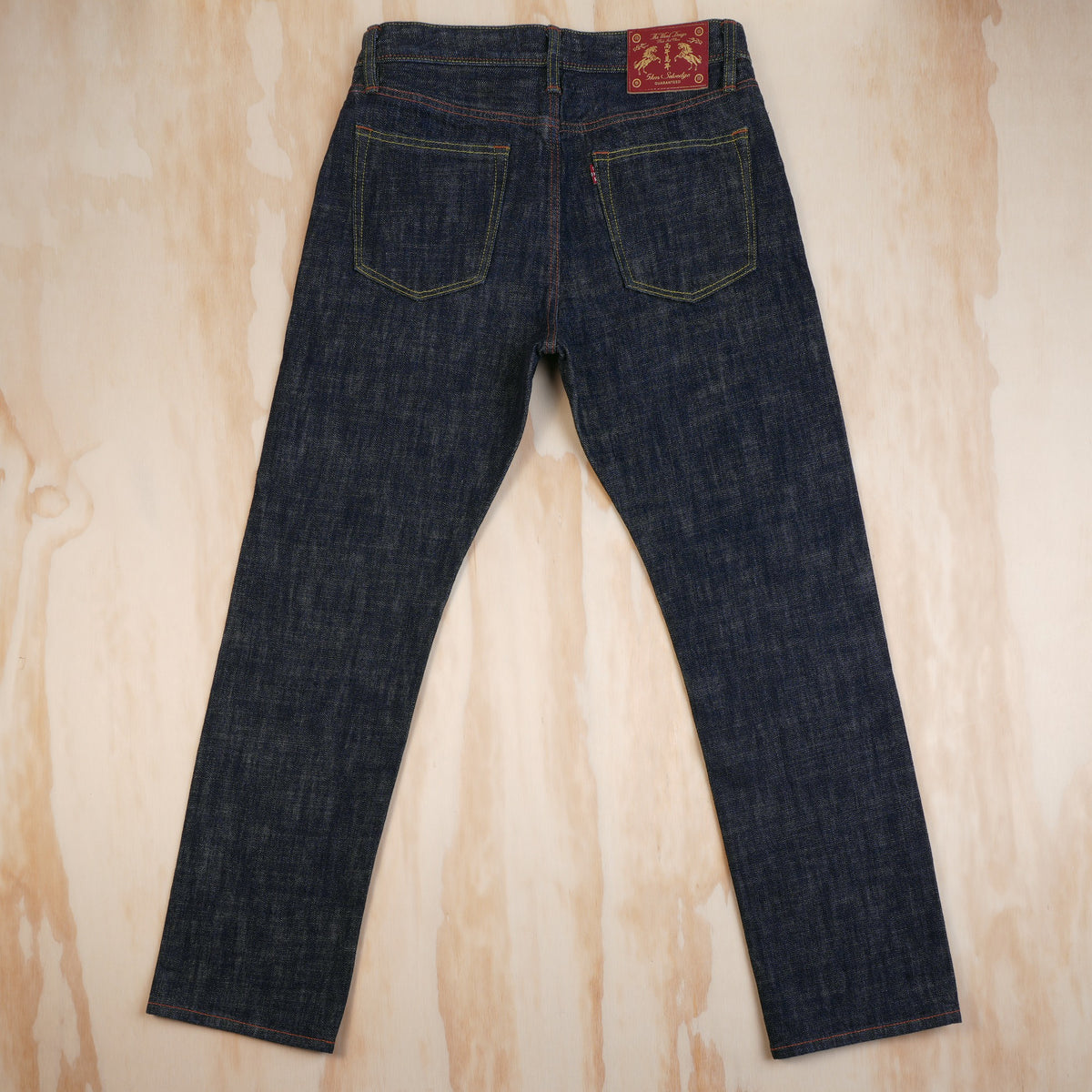 Back view of dark indigo selvedge denim jeans laid flat on a light wood background, showing the back pockets and a red brand leather patch at the waistband.