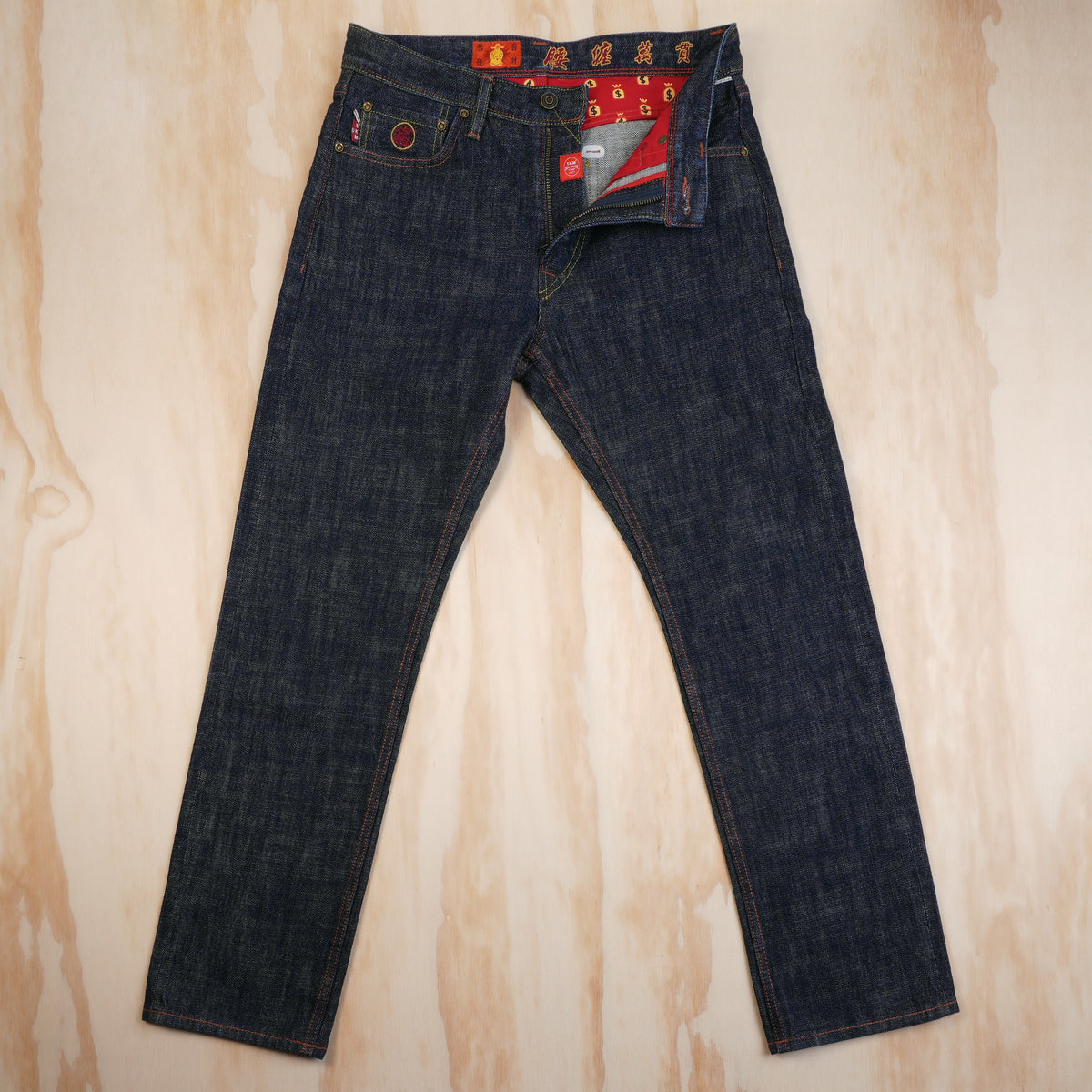 A pair of dark indigo selvedge denim jeans laid flat on a light wood-grained surface. The waistband features red and gold embroidered details with Chinese characters, and a red interior lining is visible as the fly zipper is opened.