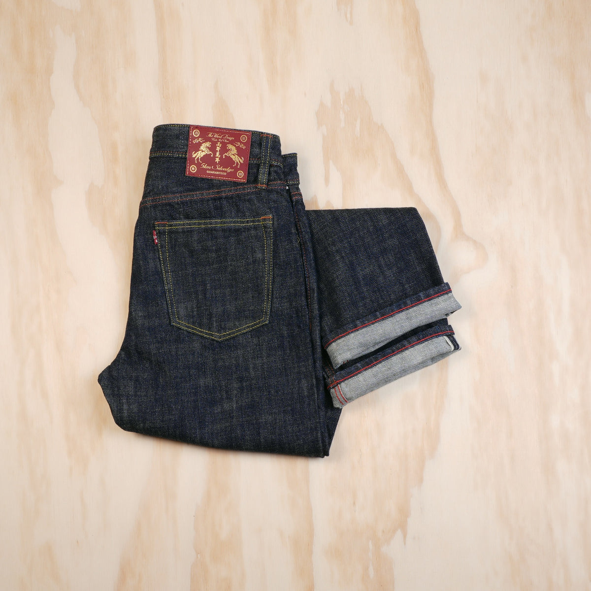 A pair of folded dark indigo selvedge denim jeans laid flat on a light wood background, with the back pocket, waistband patch and selvedge ID visible.
