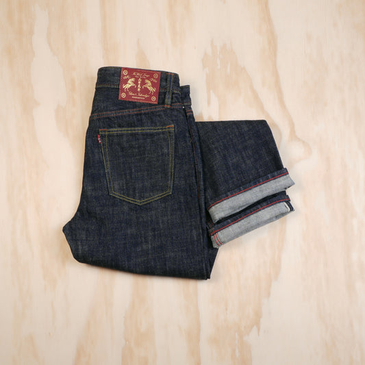 A pair of folded dark indigo selvedge denim jeans laid flat on a light wood background, with the back pocket, waistband patch and selvedge ID visible.
