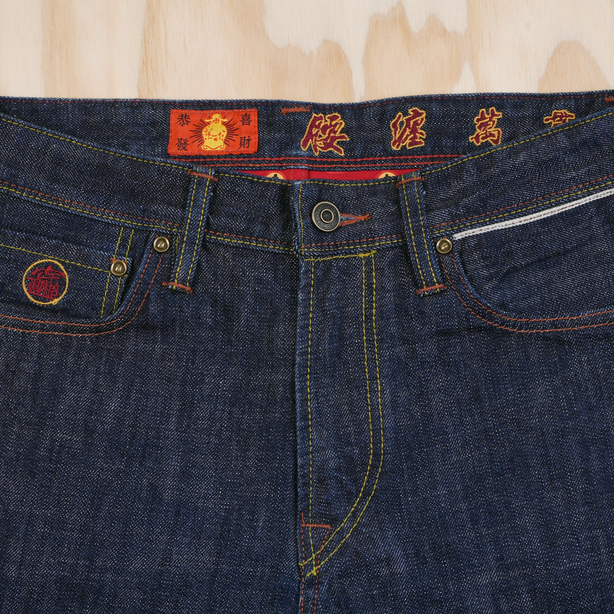 Close-up of dark indigo selvedge denim jeans laid flat on a light wood background, showing the waistband with red embroidered Chinese characters, gold-tone metal hardware, and contrast yellow stitching and selvedge ID detail above left pocket.