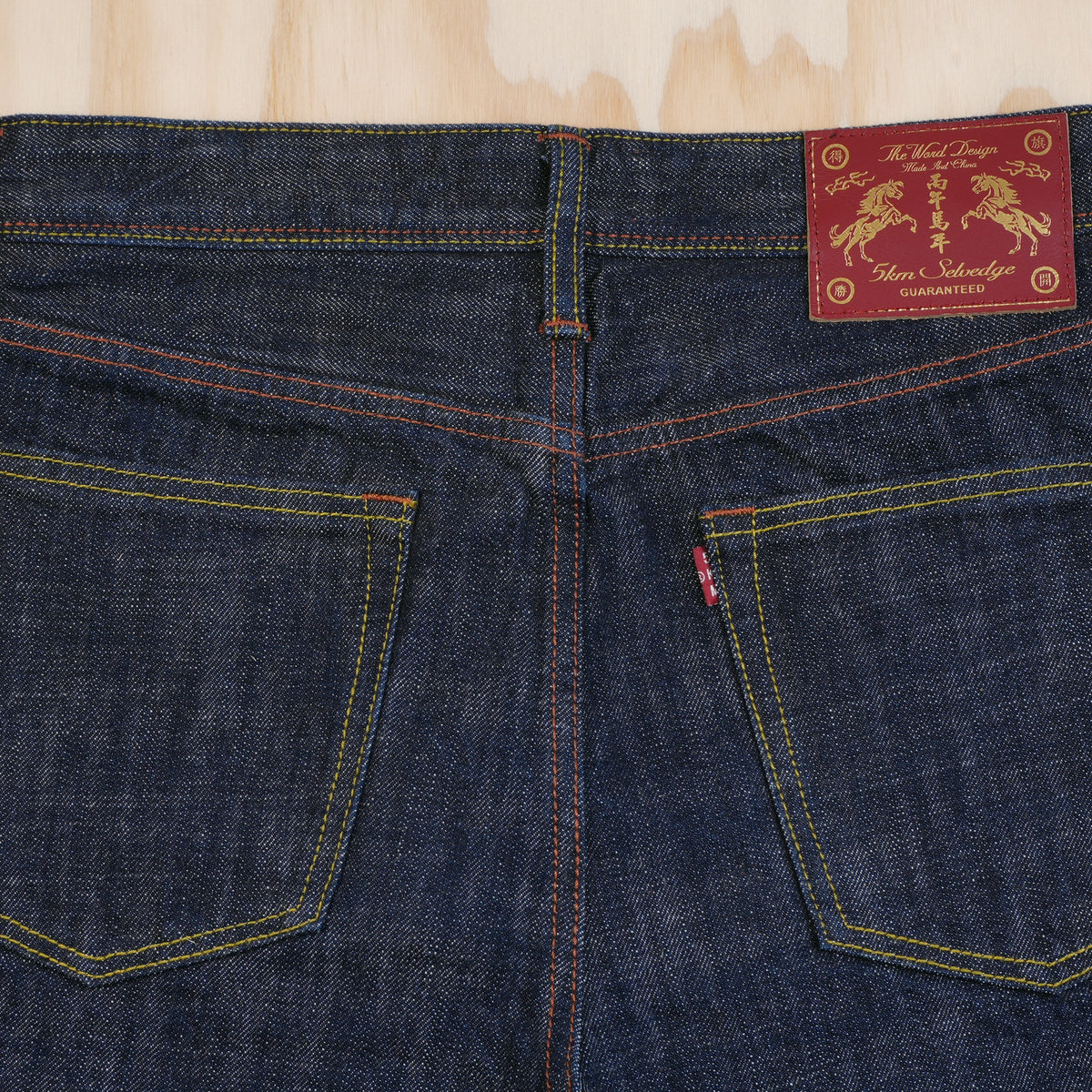 Close-up of the back waistband and pockets of dark indigo selvedge denim jeans, showing contrast yellow and orange stitching and a red leather patch with gold embroidery.