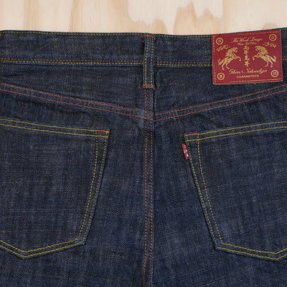 Close-up of the back waistband and pockets of dark indigo selvedge denim jeans, showing contrast yellow and orange stitching and a red leather patch with gold embroidery.