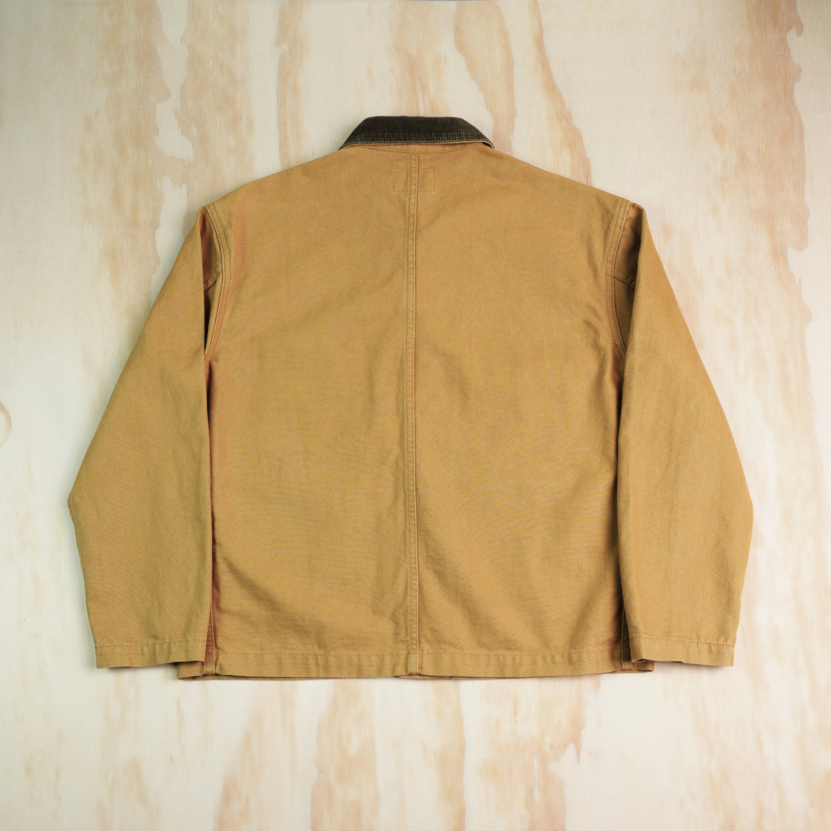 A khaki canvas chore coat shown from the back, laid flat on a light plywood background.