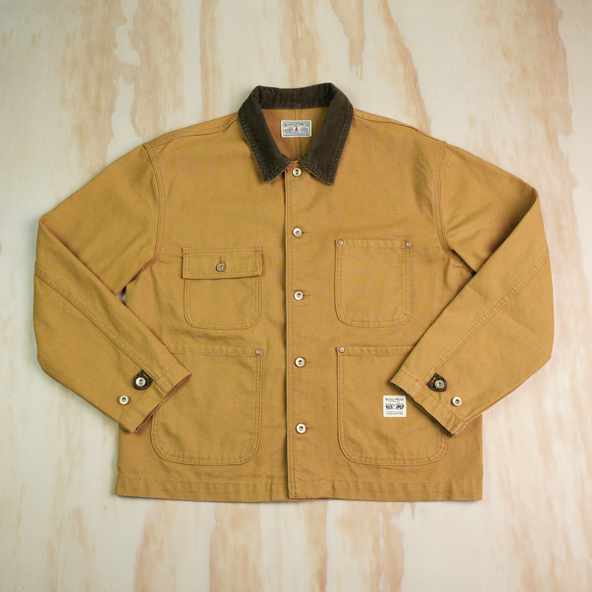 A classic khaki canvas chore coat with a dark brown corduroy collar, laid flat on a light plywood background.
