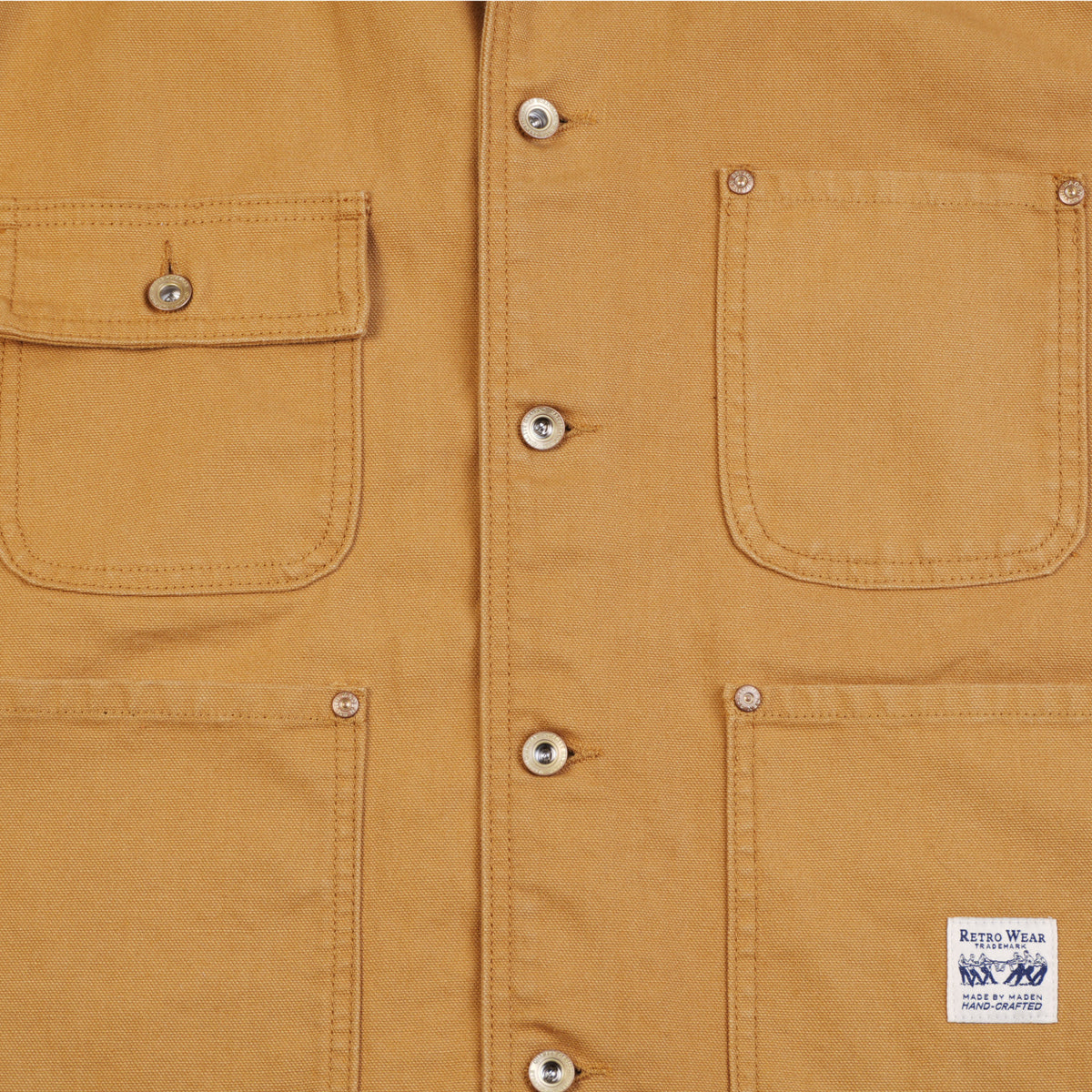 Close-up of a khaki canvas chore coat showing two front pockets with metal rivets and a white sewn-in label that reads 'RETRO WEAR MADE BY MADEN HAND-CRAFTED'.