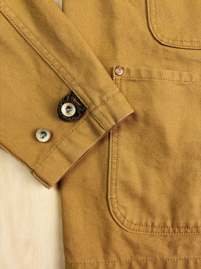 Close-up of a khaki canvas chore coat showing the sleeve cuff with metal snap buttons and a front pocket.