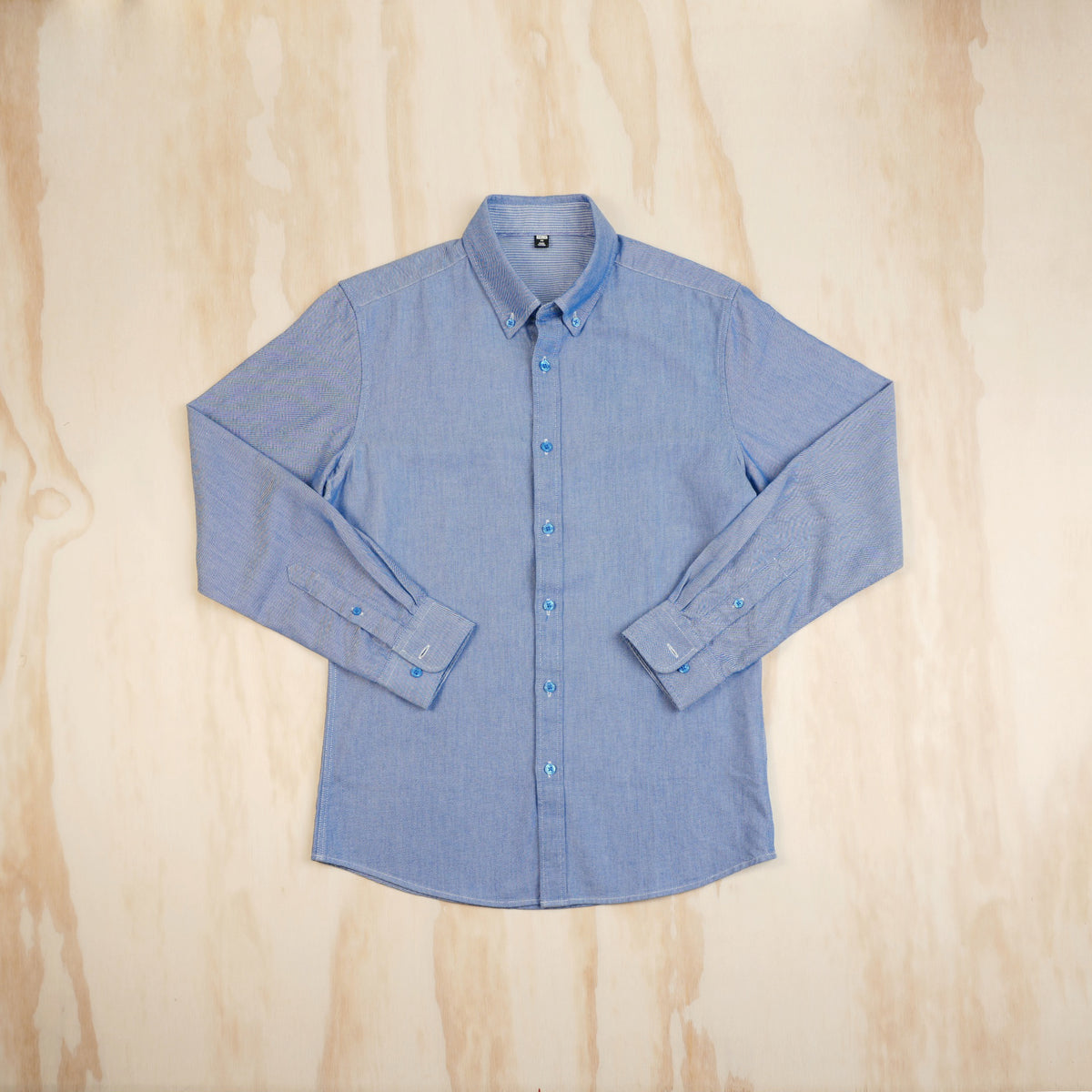A light blue long-sleeve button-up oxford cloth shirt laid flat on a light wood background, viewed from the front.
