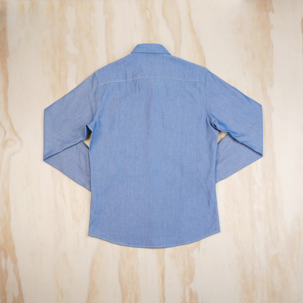 A light blue long-sleeve button-up oxford cloth shirt laid flat on a light wood background, viewed from the back.