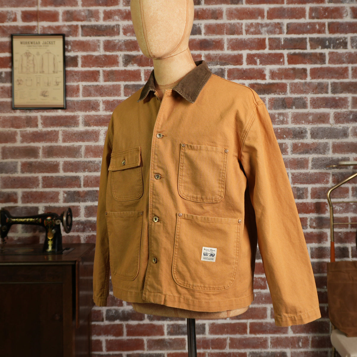 A khaki canvas utility jacket with a brown corduroy collar, displayed on a wooden mannequin torso against a red brick wall background.