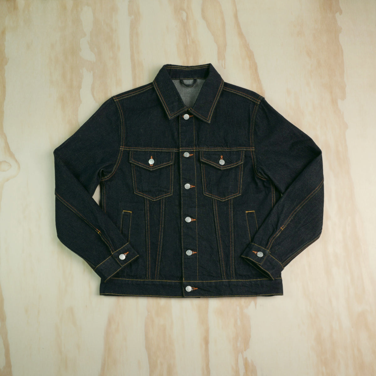 A dark indigo type 3 selvedge denim trucker jacket with contrast gold/yellow stitching, laid flat on a light wood grain background.