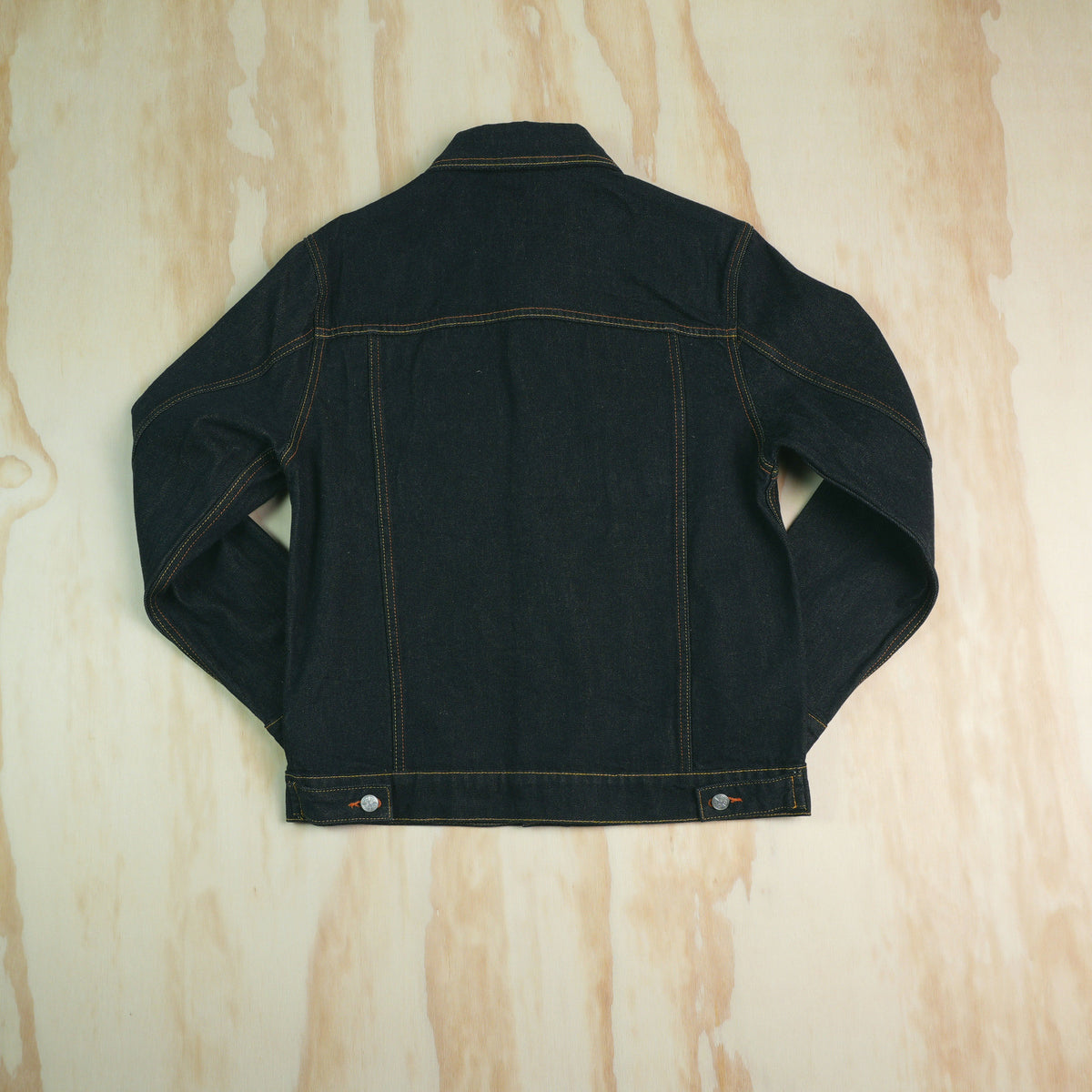 A dark indigo type 3 selvedge denim jacket shown from the back, laid flat on a light wood grain surface. The jacket features contrast orange stitching and silver metal snap buttons at the bottom hem.