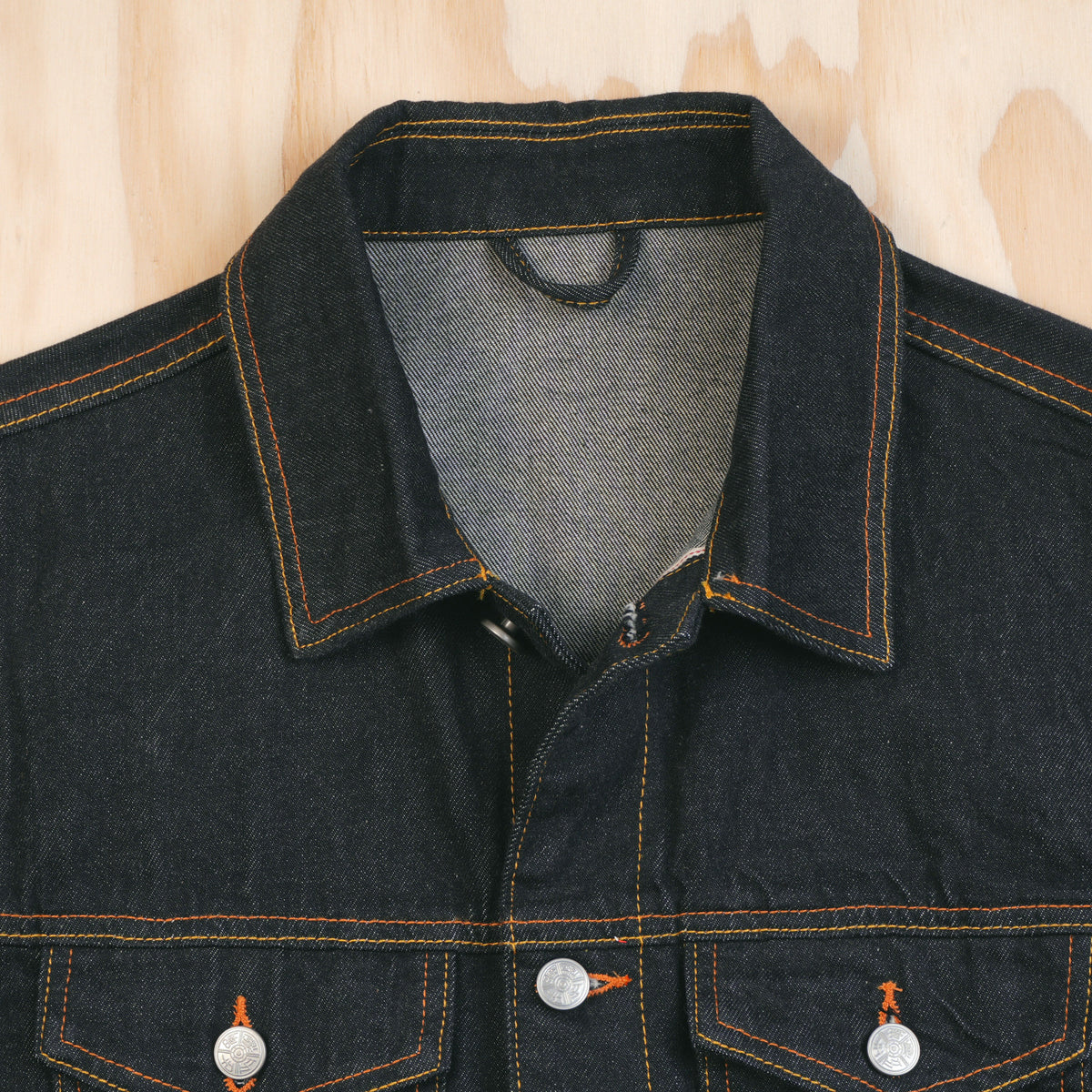 Close-up of a dark indigo type 3 selvedge denim jacket with orange contrast stitching, featuring a pointed collar, button front, and two buttoned chest pockets, laid flat on a light wood background.